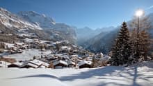 Mountain village with wooden houses, snow-covered roofs, surrounded by snowy mountains, pine trees, and a clear blue sky.