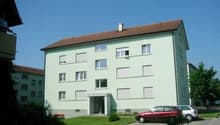 3 story apartment building, green exterior, brown roof, multiple windows, cars parked in front