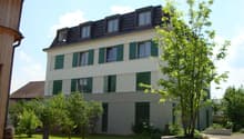 3 story apartment building, white walls, green shutters, gravel driveway, tree, plants, barrel