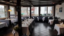 A restaurant interior with wooden beams, large windows, dark tiled floors, and tables with white tablecloths and neatly arranged wine glasses and napkins.