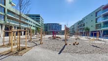 The image shows a public plaza or outdoor space surrounded by modern multi-story residential buildings. The plaza features wooden benches, sculptures, and a gravel-covered ground. The buildings have balconies visible, indicating they are likely apartments