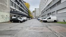 A street with parked cars along the sides, two tall buildings, concrete sidewalk, a few trees, and cloudy sky
