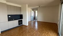 The image shows a spacious open-plan apartment with a modern kitchen and living area. The apartment features hardwood flooring throughout, white walls, and recessed lighting. There is a sliding glass door leading to what appears to be a balcony or terrace