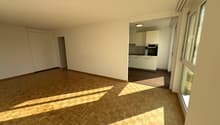 Wooden flooring, sun-drenched, large windows, empty room, white kitchen with black oven, white cabinets.