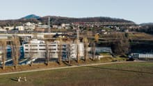 The image shows a cityscape with a mix of residential and commercial buildings, a large bridge or overpass structure, and a hilly, forested area in the background. There appears to be a parking area or lot in the foreground.
