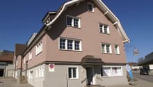 3-story building with a brown tiled roof, white trim, and multiple windows. The building has a commercial space on the ground floor and appears to be located in a town or city setting.