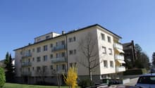 4 story apartment building, beige exterior, multiple balconies, windows, green bushes, trees, cars parked