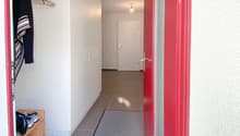 The entryway of a building with red door, tiled floors, a small carpet, a closet with clothes and shoes visible, a wooden shelf with shoes, and a hallway leading to a white door. The hallway is illuminated by a light fixture on the ceiling. The entire int