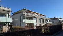 Three-story house, gray facade, glass railings on the upper floors, green roofing, covered area in the entrance, shrubs in the foreground