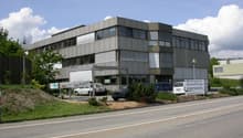 Commercial building, grey facade, large windows, cars parked in front, greenery around