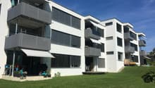 Modern white apartment building with multiple balconies, well-maintained garden, and outdoor seating area.