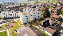 Aerial view of a residential area with multiple buildings, some with brown roofs and others with white facades, surrounded by green fields and trees.