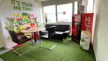 Small room with round table, two black chairs, a red sofa, a Coca Cola machine, a poster on the wall, a potted plant on the windowsill, and artificial grass flooring.