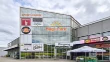 A large commercial building with a glass facade, featuring various retail and service establishments such as a sports store, an outlet store, a bistro, and a dairy and meat shop. The building is called 'Sport-Fabrik' and has a 'Hagi-Park' sign, indicating