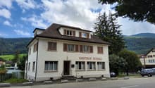 white house, brown roof, large windows, two cars parked, sign reads 'Gasthof Rossli', mountain in the background
