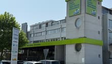 A multi-story commercial building with a Steinmann cafe and restaurant on the ground floor. The building has a large green and white sign advertising their cafe, lunch, take away, and chocolate offerings. There are several cars parked in front of the buil