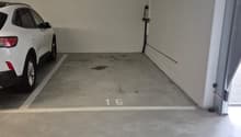 The image shows a garage space with a white SUV parked inside. The floor is concrete and there is a number '15' painted on the floor, indicating this is parking spot number 15. The walls are plain white and there are some utility pipes or wires visible on