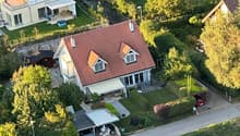 House with red roof, car parked in garage, surrounded by trees and grass, aerial view