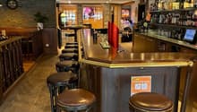 bar counter, stools, wooden flooring, wooden bar, bartop, shelves with alcohol bottles, gaming machines, plants