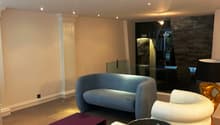 Spacious living room with modern furniture, including a blue sofa, white and beige armchairs, and a purple coffee table on wheels. The room has recessed lighting in the ceiling and a stone accent wall with a fireplace.