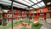 Large open interior space with a grass lawn, wooden seating area, tables, and staircases; orange and red walls with glass doors and windows.