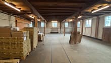 Empty warehouse with wooden beams, fluorescent lights, wooden pallets, stacks of boxes, concrete floor, windows