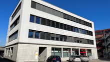 A multi-story commercial building with a modern, minimalist design. The building has large windows and a concrete exterior. There is a parking area in front of the building with several cars parked.
