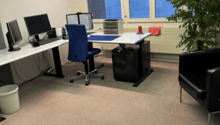 The image shows an office space with a desk, computer, and other office equipment. There is a large window with a view of what appears to be a balcony or terrace outside.