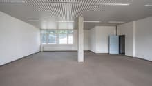 Large open office space with high ceilings, exposed beams, and large windows. The room has a concrete column in the center and a refrigerator in the corner. The space appears to be unfinished and ready for customization.