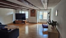 Spacious living room with hardwood floors, exposed wooden beams on the ceiling, a large TV mounted on the wall, a sofa, and a coffee table. There is also a dart board on the wall and a potted plant in the corner.