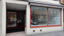 The image shows the exterior of a commercial building with a storefront. The building has large windows with red frames, and the entrance is through a doorway. There are cars parked on the street in front of the building.