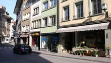 A street with buildings on both sides, a car driving, and a van parked on the side. The buildings have glass windows and balconies. The sidewalk has potted plants and a shop with a white awning. The shop has glass windows with potted plants and flowers di