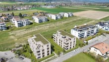 The image shows a residential area with several multi-story apartment buildings surrounded by green fields and hills in the background. The buildings have balconies and there are parking spaces visible in the foreground.