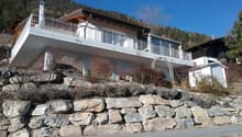Two-story house, white exterior, balcony with railings, stone retaining wall, mountains in the background.