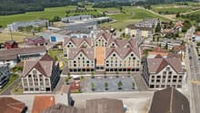 The image shows an aerial view of a town with a mix of residential and commercial buildings. The town has a central square or plaza surrounded by multi-story buildings with red tile roofs. There are also some industrial or warehouse buildings in the backg