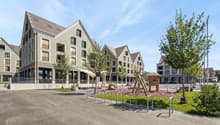 The image shows a residential area with several multi-story apartment buildings. The buildings have a modern, uniform architectural style with gabled roofs and large windows. There is a public plaza or square in the foreground with benches, bike racks, an