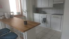 Open kitchen with a wooden island, white cabinets, white tiled floor, a window with shutters, a sink with faucet, potted plant, a kettle, toaster, and a cooking stove.