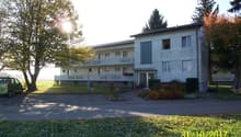 2 story white building, multiple windows, balcony, parking lot, trees, plants, green car