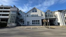 A multi-story commercial or office building with a modern, glass-heavy architectural design. The building has a large central structure with several wings and annexes. The exterior is primarily composed of light-colored walls and large windows. There is a
