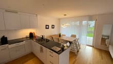 modern kitchen with white cabinets, black countertops, island with sink, dining table, chairs, wooden floor, sliding glass door to patio, pendant light, mirror, framed pictures