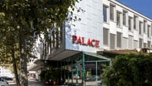 2-story building, white exterior, large glass windows, sign 'PALACE', plants, tree, sidewalk, cars