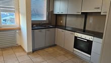 Kitchen with tiled flooring, a sink with faucet, a black countertop, built-in appliances including an oven and a dishwasher, white cabinets, and windows with blinds.