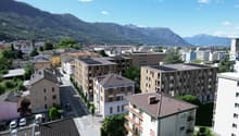 Aerial view of a town with several buildings, mountains in the background, solar panels on some buildings, cars parked on the street, trees and greenery