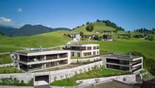 Modern architectural design, multi-story building, white exterior, large windows, solar panels on the roofs, located on a hillside with green surroundings, stone walls, and gardens.