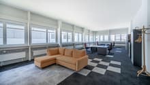 Large office space with a corner sofa, desk, and office chairs, equipped with white radiators and large windows.