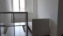 Minimalist bedroom with white walls, hardwood flooring, a metal shelving unit, and a bed frame. The room appears to be in a state of renovation or furnishing.