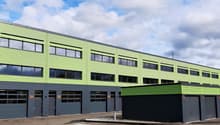 Large green and grey industrial building, several large windows, roll up garage doors on the ground floor, three garages on the side