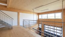The image shows a large open space with a wooden ceiling and walls. There is a metal railing along the second floor balcony, and the floor is made of wood planks. The room has large windows that provide natural light. The overall design appears to be mode
