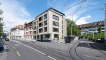 Multistory residential building, modern design, several windows, elevated ground floor, street tram tracks, bus stop, trees, parked cars
