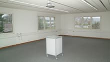 Large open room with white walls, ceiling-mounted projector, and multiple windows providing natural light. The room has a carpeted floor and a mobile cart or cabinet in the center.